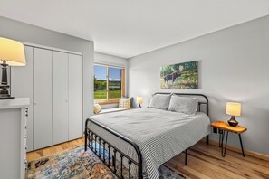 A bedroom with a bed, bedside lamps, a window with a view, a painting on the wall, and a wardrobe.