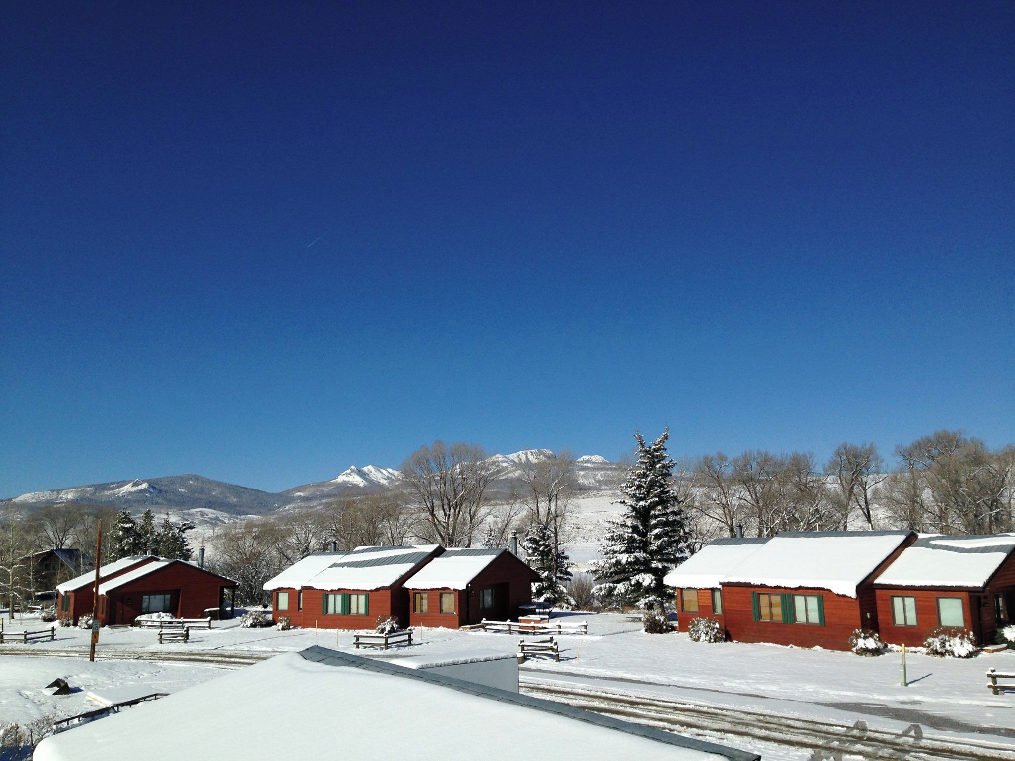 A row of red cabins with snow-covered roofs, set against a backdrop of mountains and clear blue sky.