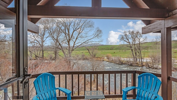 Een overdekt terras met twee blauwe Adirondack-stoelen en een tafeltje, met uitzicht op een rivier en een begroeiend landschap met bomen.