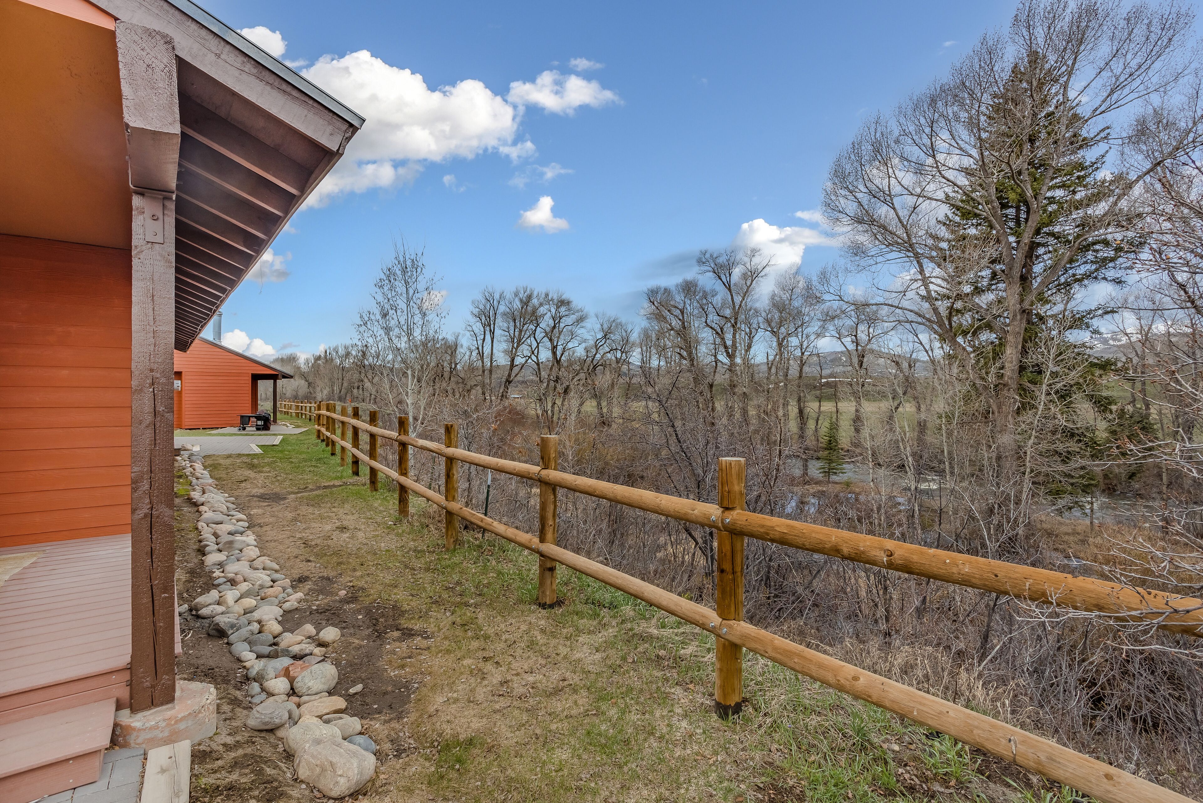 A wooden deck leading to a building with a sloped roof, surrounded by a wooden fence and a natural landscape with trees and a clear sky.