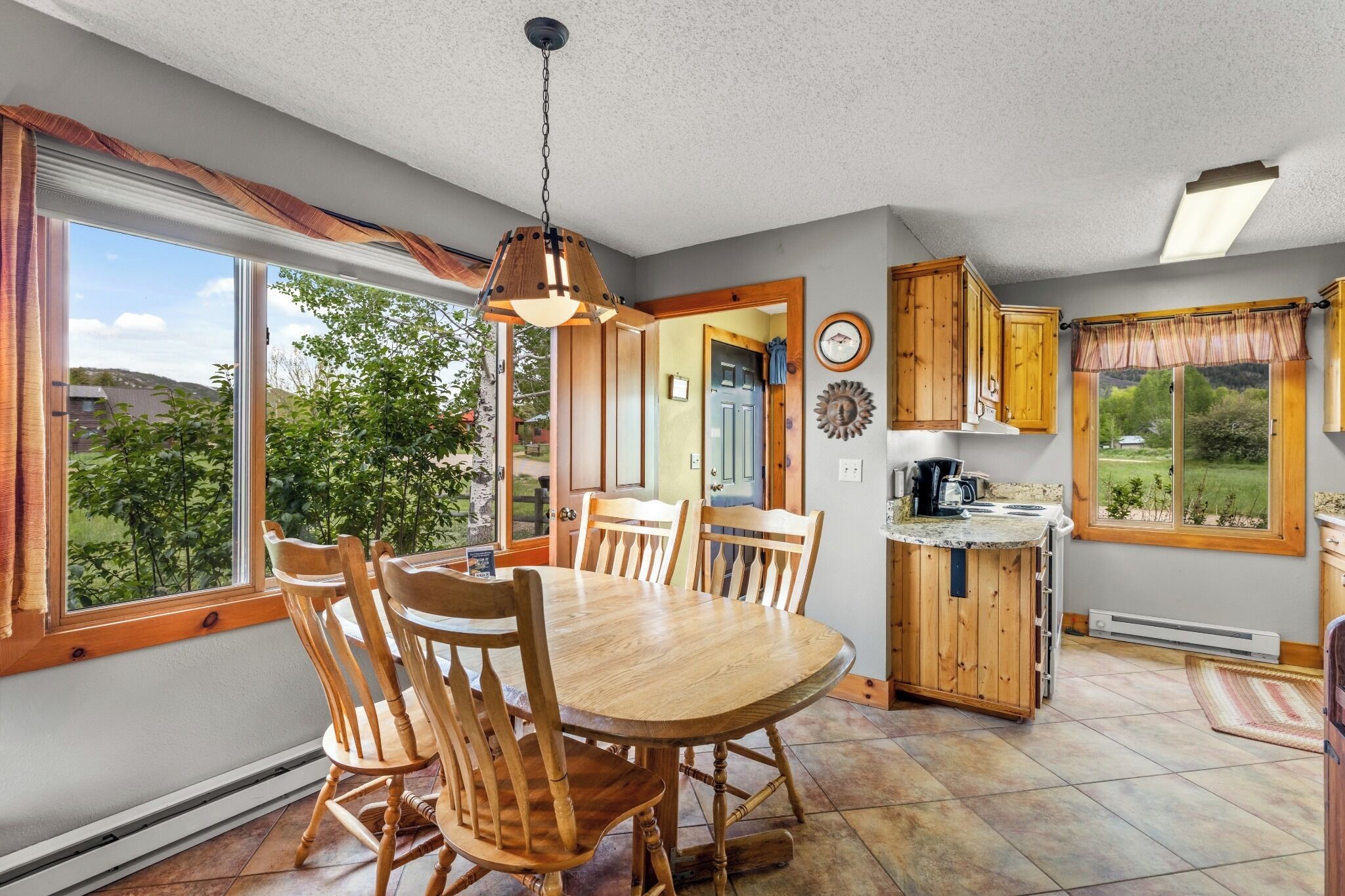 A dining area with a wooden table and chairs, a pendant light, and a view of greenery outside.