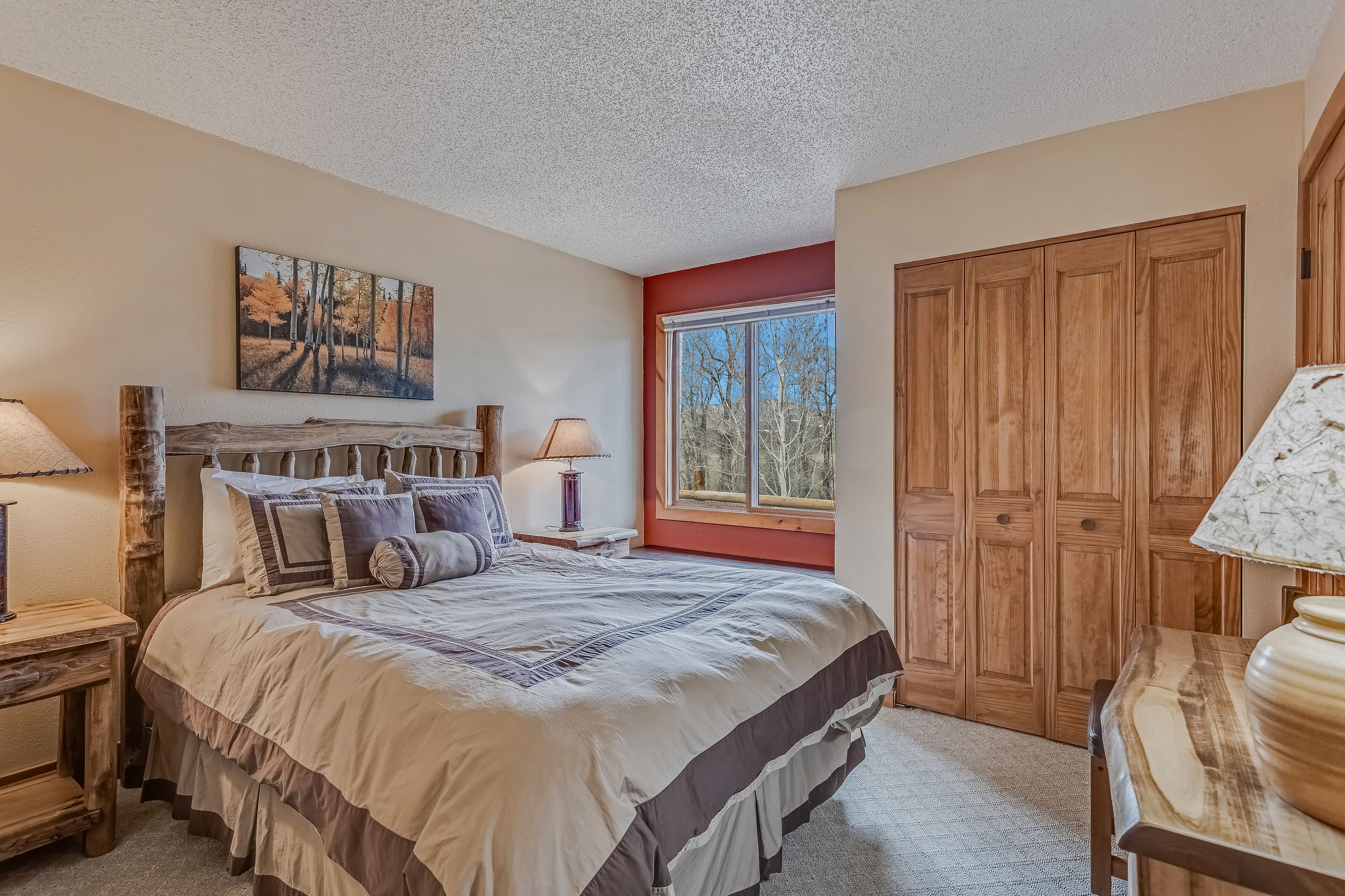 A bedroom with a large bed, wooden headboard, a window with a view, a wooden wardrobe, and a painting on the wall.