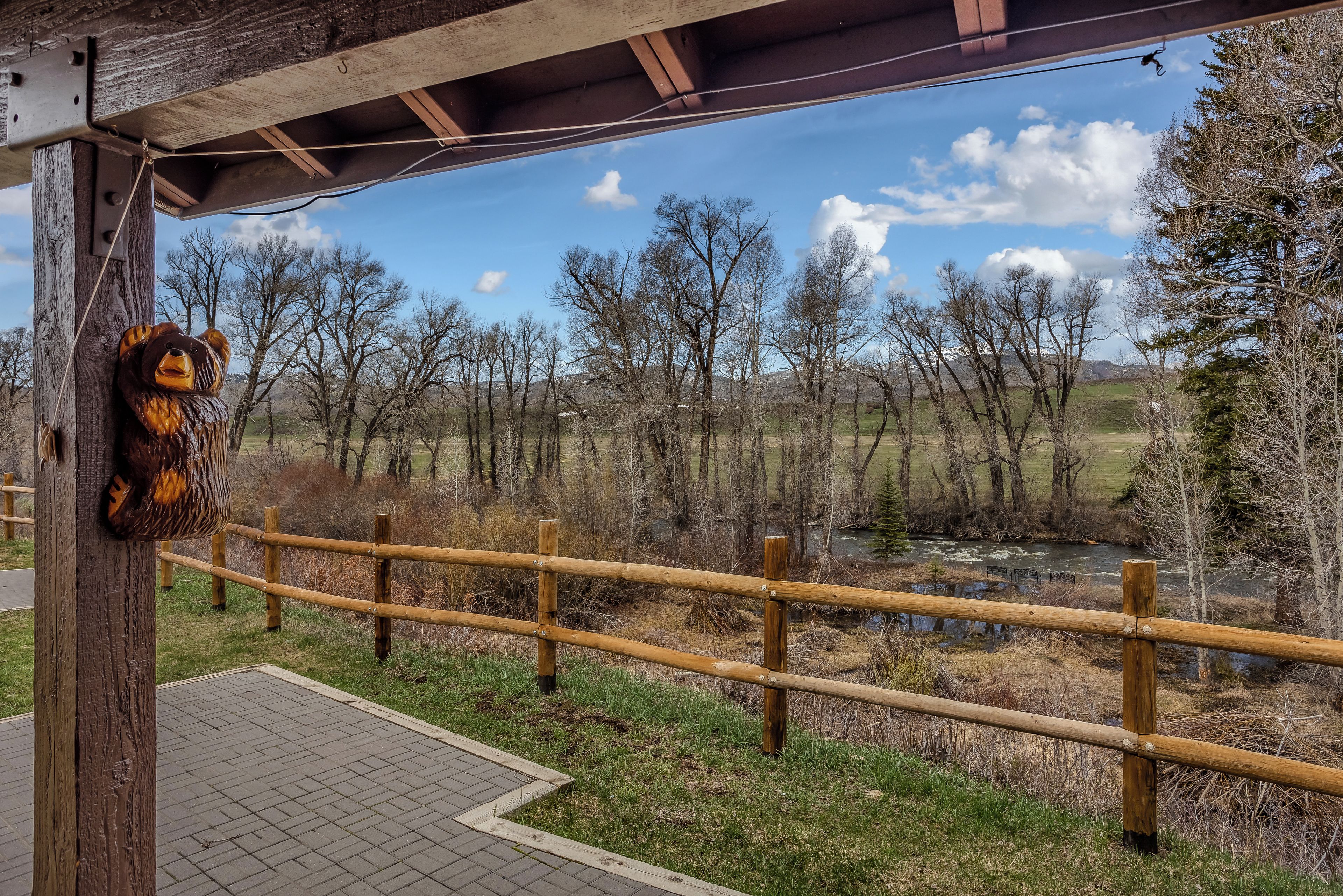 A wooden fence with a bear sculpture on a post, overlooking a river and trees.