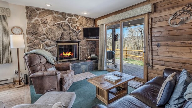 A cozy living room with a stone wall, fireplace, leather sofas, and a large window overlooking a deck. - Grizzly - Stunning mountain views on the Elk River (Clark)