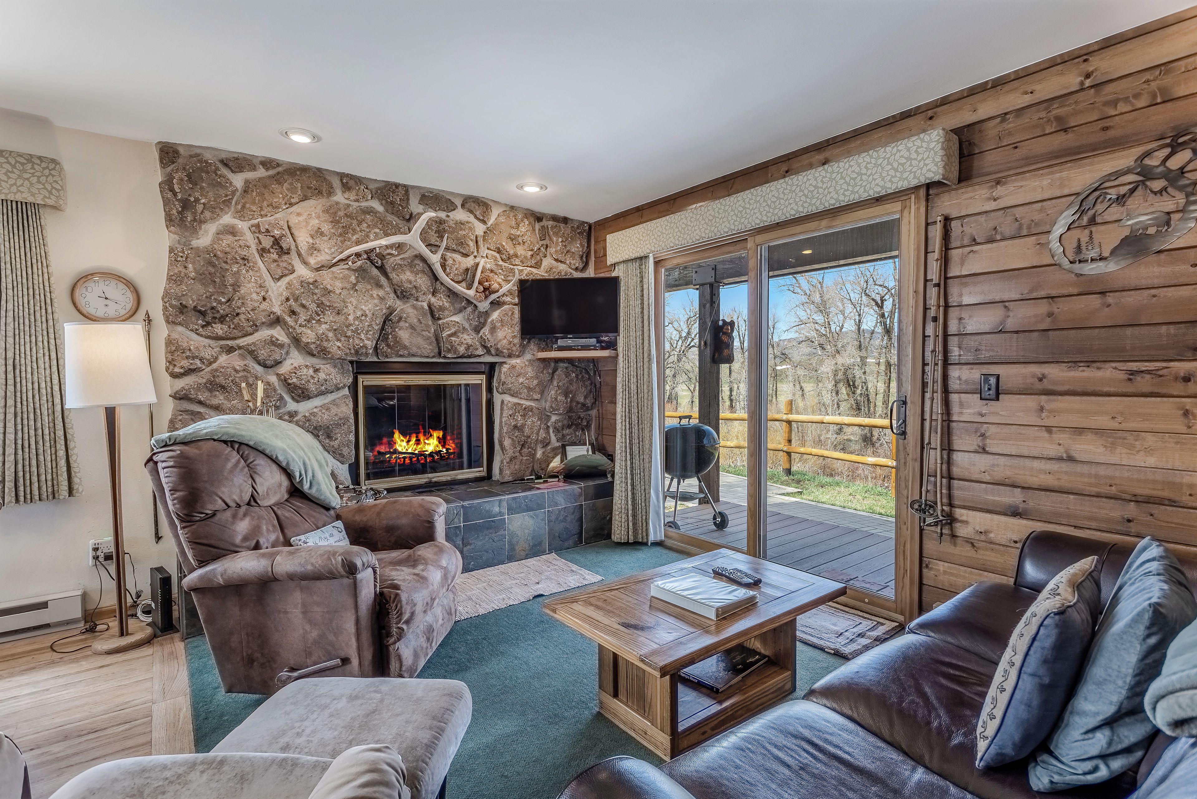A cozy living room with a stone wall, fireplace, leather sofas, and a large window overlooking a deck.