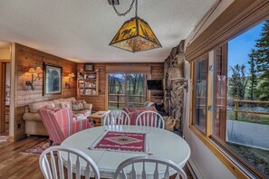 L’intérieur chaleureux d’un chalet avec une table à manger ronde, des chaises blanches, un plafond en bois et une grande fenêtre donnant sur le paysage extérieur.