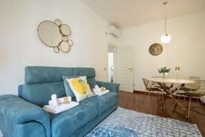 A living room with a blue sofa, a round mirror, a dining table with chairs, and a clock on the wall.