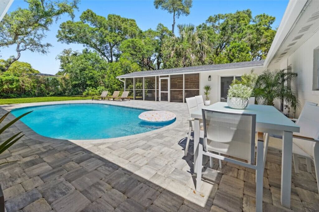 Ein Schwimmbecken mit einer Steindeck, eine überdachte Terrasse mit Sitzgelegenheiten und üppiges Grün im Hintergrund.