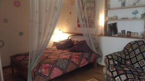 Un dormitori amb un llit, una taula de nit, una làmpada, una cadira i una estantàpia amb objectes decoratius.
