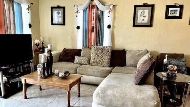 A living room with a beige sofa, a wooden coffee table, and a television. There are framed pictures on the wall and a curtain with a colorful pattern. - Comfortable Home in Hallandale Beach (Hallandale beach)