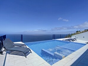 A modern infinity pool with a clear view of the ocean, accompanied by lounge chairs and a small hot tub.