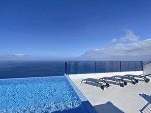 A clear blue sky, a swimming pool, and lounge chairs overlooking the ocean.