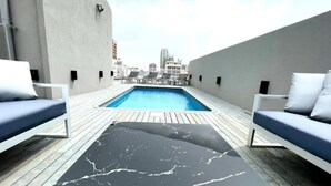 Un área de piscina en la azotea con diseño moderno, que incluye una mesa de mármol negro y dos sillones de descanso azules.
