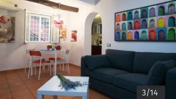 A living room with a dark green sofa, a white coffee table with a bouquet, a dining table with chairs, and a colorful wall mural. - Vaticanhouse  (Roma)