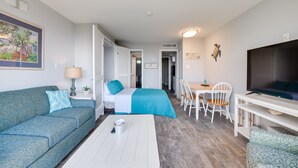 A modern hotel room with a sofa, a bed with a blue bedspread, a dining area with chairs, and a TV on a stand. - Myrtle Beach Condo w/ Ocean View + Pool Access! (Myrtle Beach)