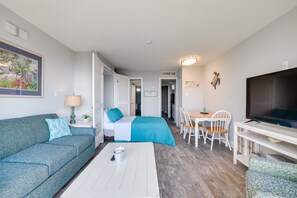 A modern hotel room with a sofa, a bed with a blue bedspread, a dining area with chairs, and a TV on a stand.