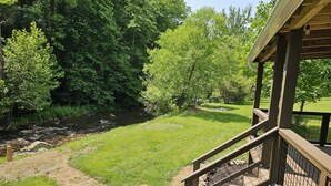 Une terrasse en bois avec une rambarde en métal, donnant sur une zone herbeuse, une rivière et une forêt dense.