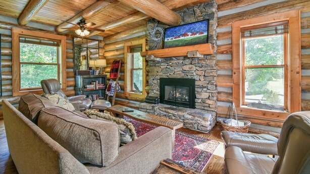 A log cabin interior with a stone fireplace, leather furniture, and large windows.