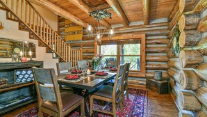A log cabin interior with a wooden dining table set for four, surrounded by chairs. A staircase with wooden railings leads to the upper level.