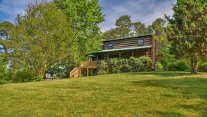 A log cabin with a deck and a covered porch, surrounded by lush greenery and trees.