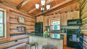 A log cabin kitchen with wooden cabinets, a black refrigerator, a microwave, and a sink. There are two stools and a clock on the wall.