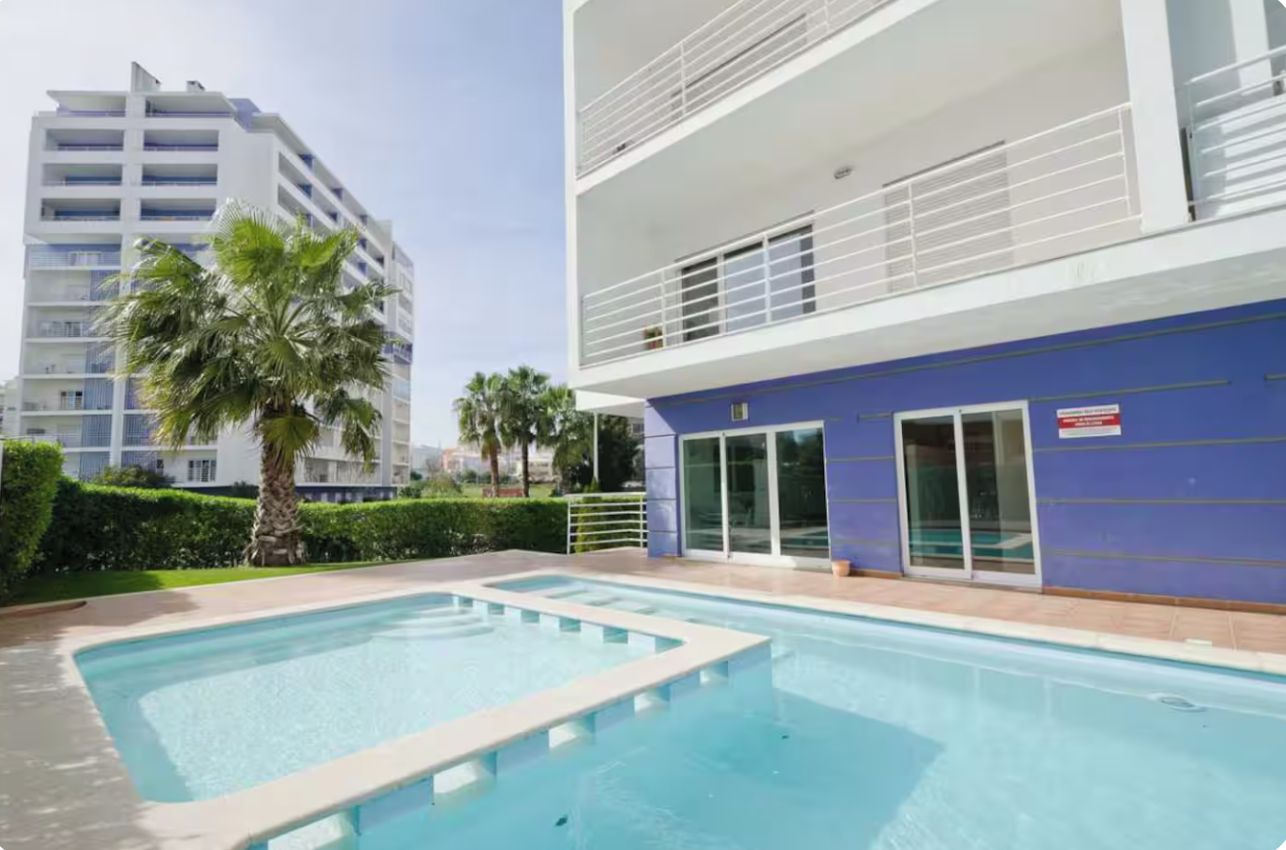 Un espace piscine avec un bâtiment moderne, un palmier et un ciel dégagé.