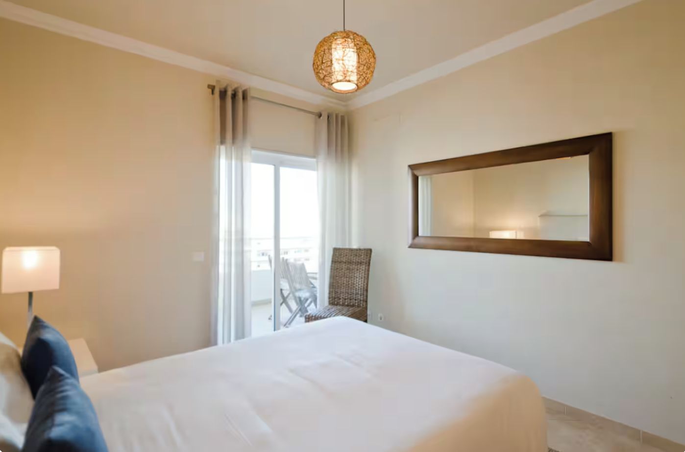 A hotel room with a bed, a chair, a lamp, a large window, and a mirror on the wall.