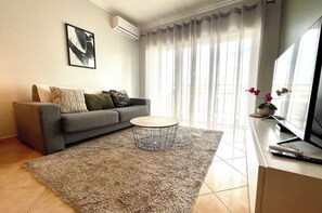A living room with a grey sofa, a round coffee table, a rug, a TV on a stand, and sheer curtains. - One-bedroom apartment with a pool, air conditioning, and a balcony with a grill, near Praia da Rocha (Portimão)
