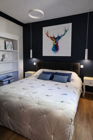 A bedroom with a large bed, two bedside tables, a white shelving unit, and a colorful abstract painting on the wall.