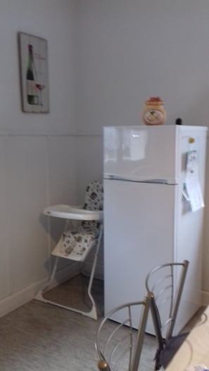 A small kitchen with a white refrigerator, a white table with a patterned tablecloth, and a chair with a metal frame.