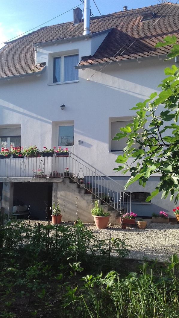 A two-story building with a tiled roof, a balcony with potted plants, and a staircase leading to the entrance.