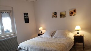 Un dormitorio con cama, dos mesitas de noche con lámparas, un radiador, una ventana con persianas y tres cuadros en la pared.