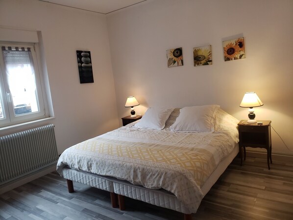 A bedroom with a bed, two bedside tables with lamps, a radiator, a window with blinds, and three wall-mounted pictures.