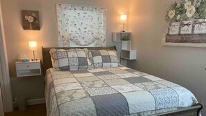 A bedroom with a bed, two bedside tables, a nightstand, a wall-mounted shelf, and a floral arrangement. - Country View Cottage-Near Massanutten, Skyline drive,&JMU.A home away from home! (Elkton)