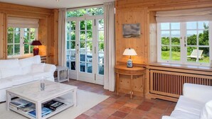 A spacious living room with a white sofa, a coffee table with books, a lamp, and a view of the outdoors through large windows.
