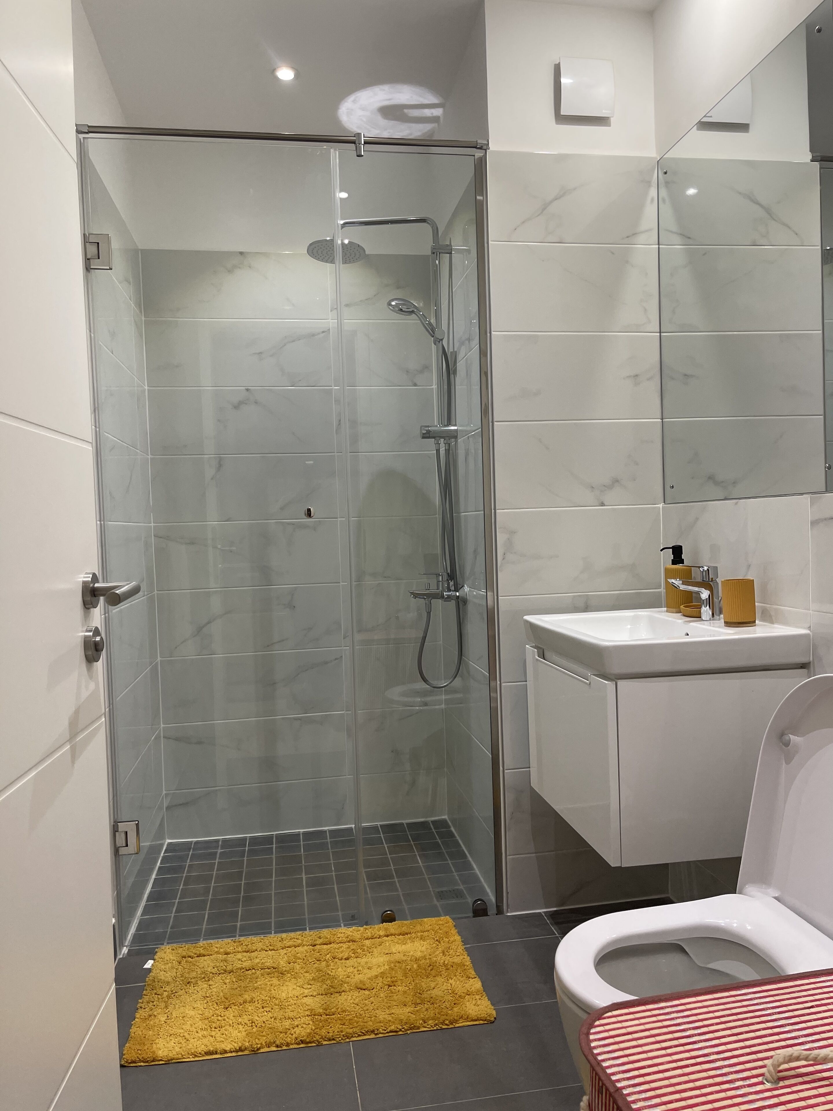 A modern bathroom with a glass-enclosed shower, a white sink with a mirror above, and a yellow rug on the floor.