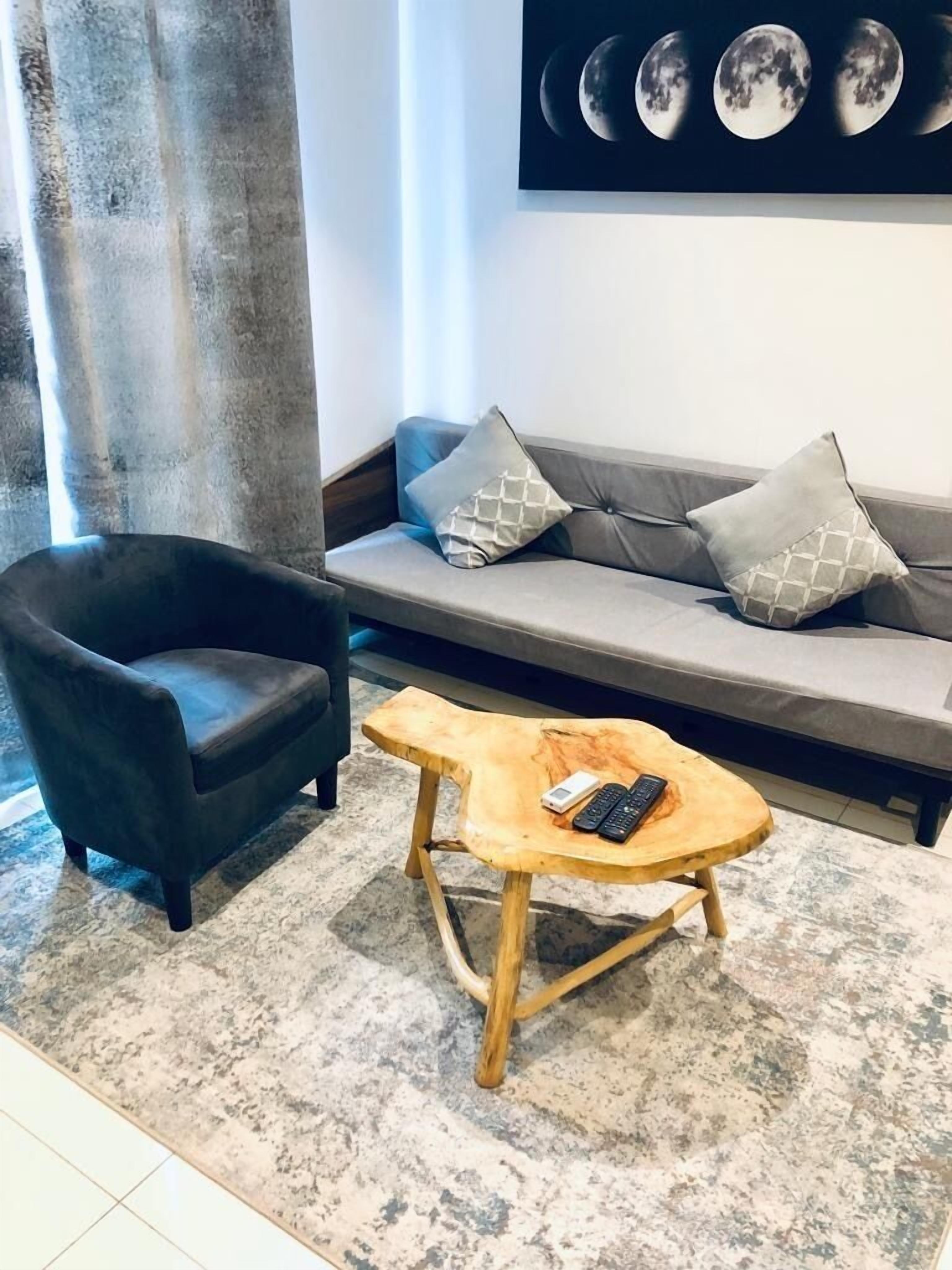 A living room with a dark armchair, a wooden coffee table with remotes, a grey sofa with patterned cushions, and a wall-mounted moon phase display.