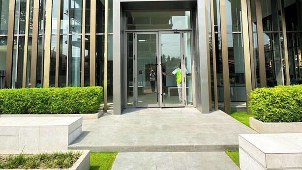 Un edificio moderno con entrada de vidrio, listones verticales de madera y un área ajardinada con césped y arbustos.
