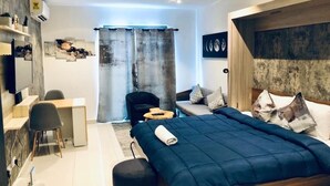 Un dormitorio moderno con una cama grande, un escritorio y una silla. Hay una cortina, un estante en la pared y un cuadro.