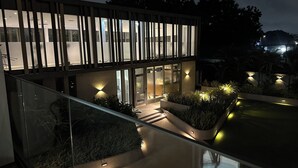 Un edificio moderno con amplios ventanales y una escalera de cristal. Hay plantas e iluminación exterior.