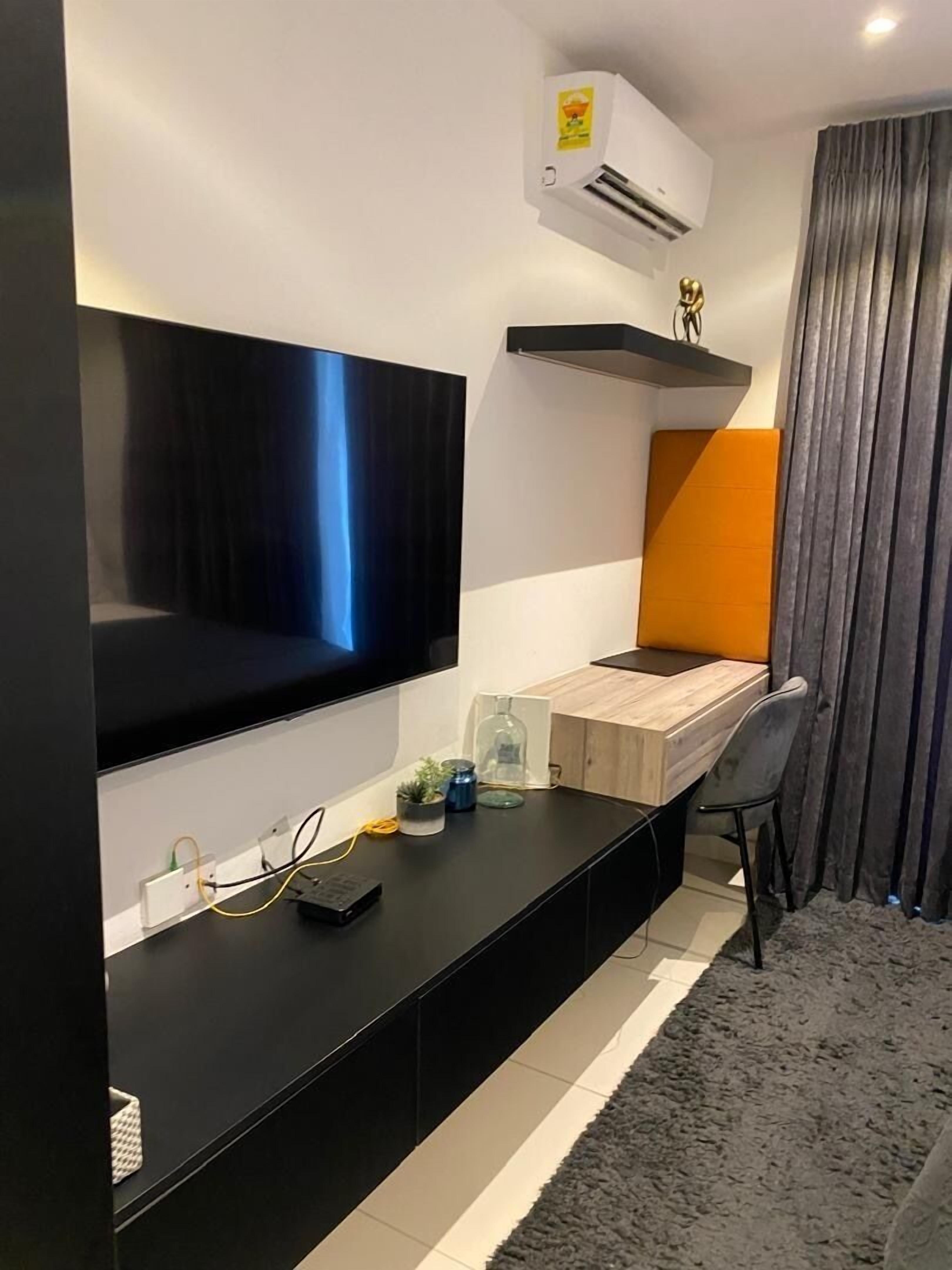 A modern living room with a flat-screen TV mounted on the wall, a wooden desk, a chair, and a grey rug.
