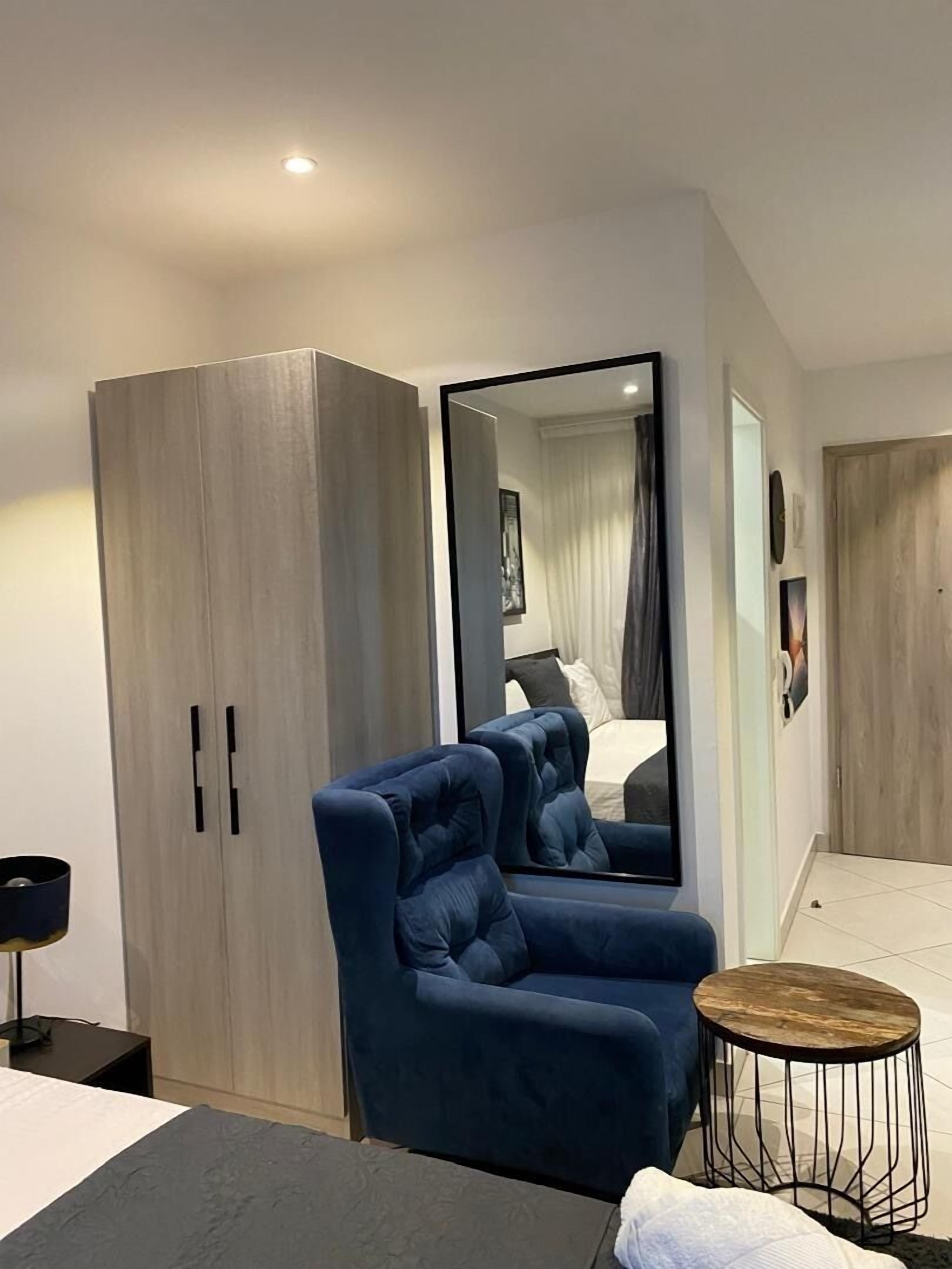 A modern bedroom with a large wardrobe, a blue armchair, a round wooden side table, and a bed with a white duvet.