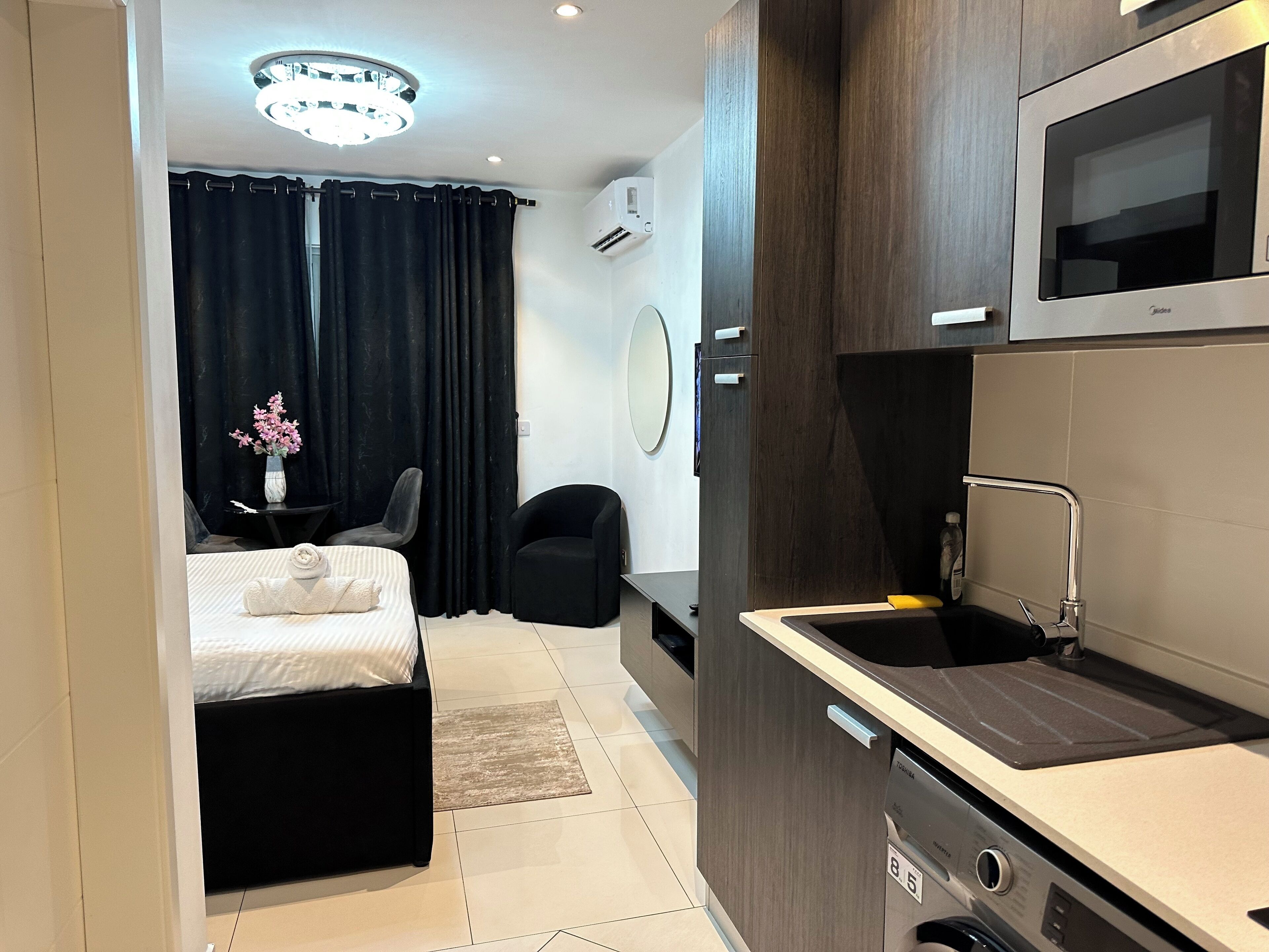 A compact hotel room with a bed, a small desk, a chair, a kitchenette, and a bathroom.