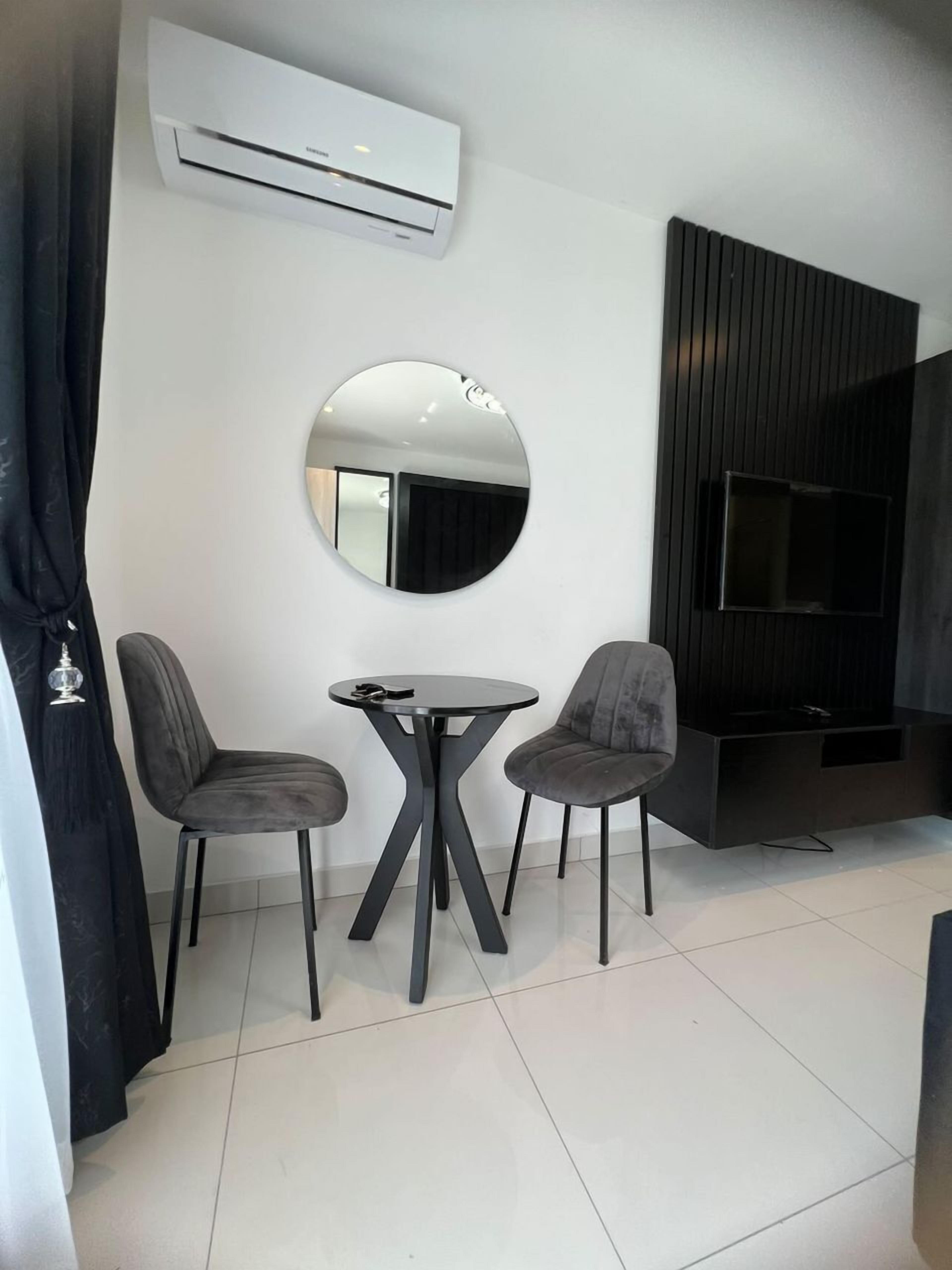 A modern living room with a round mirror, two grey chairs, a small round table, a flat-screen TV, and a built-in entertainment unit.