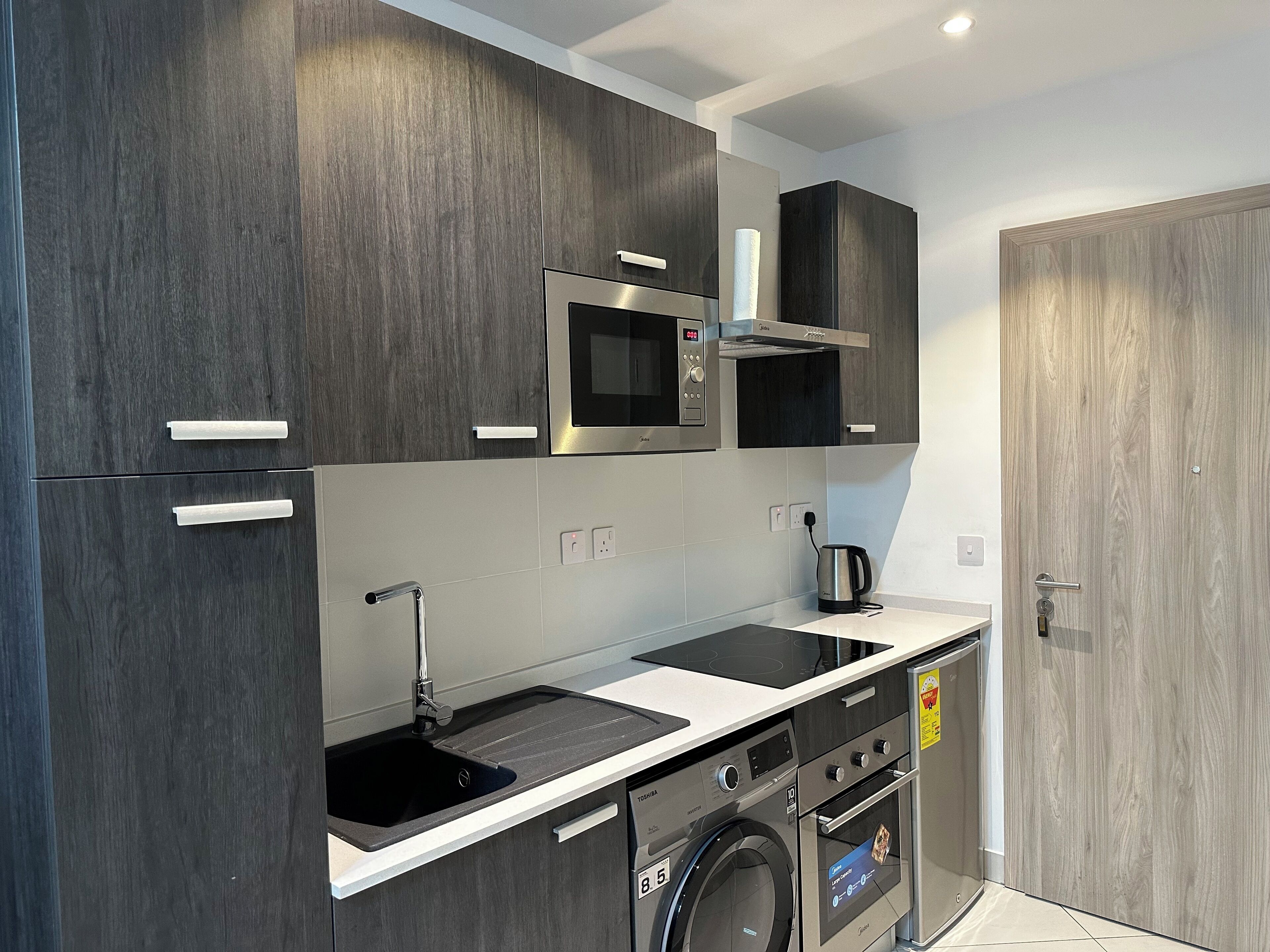 A modern kitchen with dark wood cabinets, a stainless steel microwave, a built-in oven, and a washing machine.