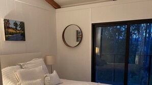 A bedroom with a large bed, a round mirror, a painting, and a view of trees through a glass door.