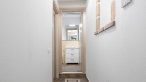 Un couloir étroit, doté d’un plancher en bois, mène à une salle de bain avec un lavabo blanc et un mur recouvert de carreaux.