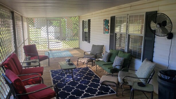 A covered patio area with a wooden floor, red and green chairs, a small table, and a blue patterned rug.