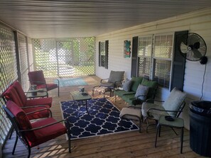 A covered patio area with a wooden floor, red and green chairs, a small table, and a blue patterned rug.