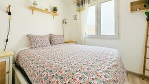 A bedroom with a floral bedspread, a hanging wicker light fixture, a window with curtains, and a wooden shelf with plants. - Renovated house and tastefully decorated in La Chaume (Les Sables-d´Olonne)